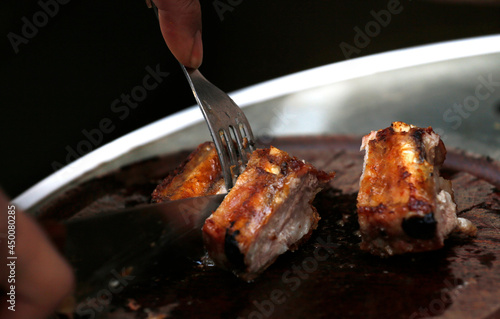 Pork ribs. Person cutting roast beef with knife and fork. Tasty roast. Black background. Fork with portion of pork or beef. Food. Barbecue. 