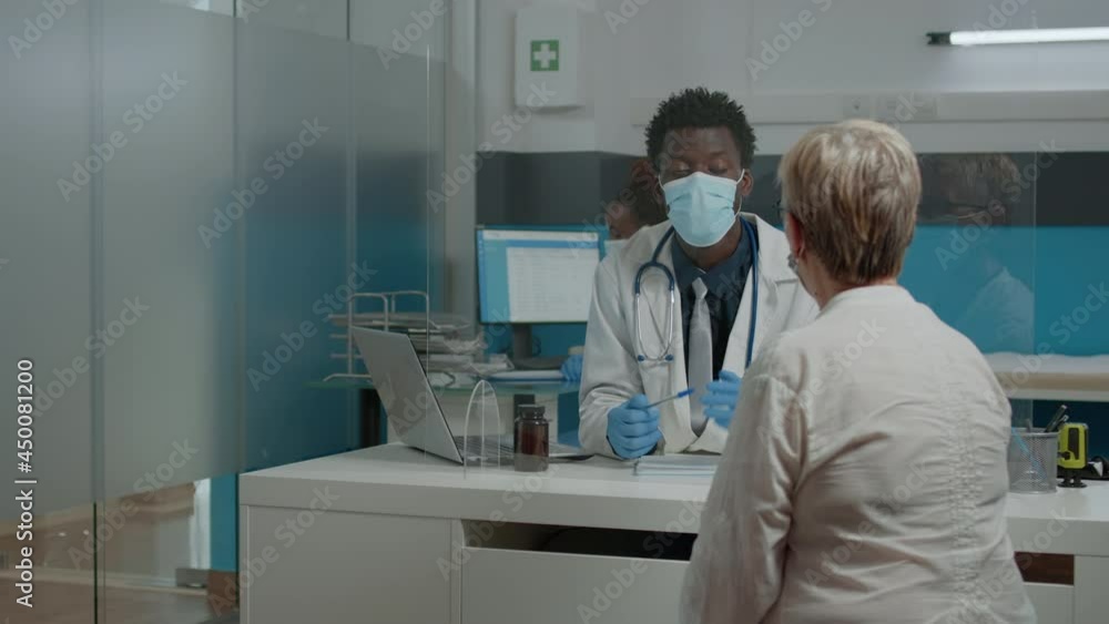 Young medic wearing face mask while consulting elder patient behind plexiglass wall at medical facility. Doctor helping senior woman with healing treatment for healthcare during pandemic Young medic wearing face mask while consulting elder patient behind plexiglass wall at medical facility. Doctor helping senior woman with healing treatment for healthcare during pandemic