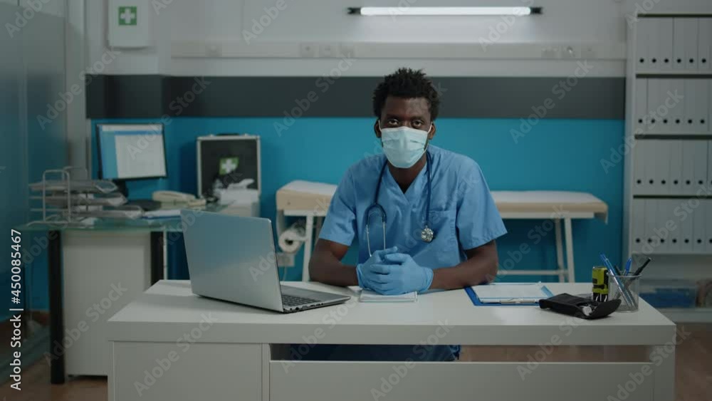 Portrait of young person working as nurse in cabinet at healthcare facility. Medical assistant sitting at white desk looking at camera with face mask, gloves, uniform and stethoscope Portrait of young person working as nurse in cabinet at healthcare facility. Medical assistant sitting at white desk looking at camera with face mask, gloves, uniform and stethoscope