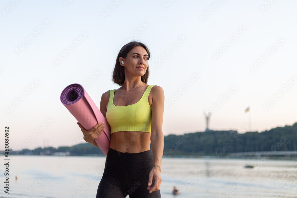 Good looking in perfect shape slender woman in sport wear at sunset on city beach with yoga mat and wireless headphones strong abs, sport body motivation Good looking in perfect shape slender woman in sport wear at sunset on city beach with yoga mat and wireless headphones strong abs, sport body motivation