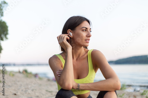 Good looking in perfect shape slender woman in sport wear at sunset on city beach resting aafter workout listening music in wireless headphones