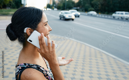 Adult woman with a hearing impairment uses a hearing aid in everyday life, talking on phone in urban city outdoor. Hearing solutions