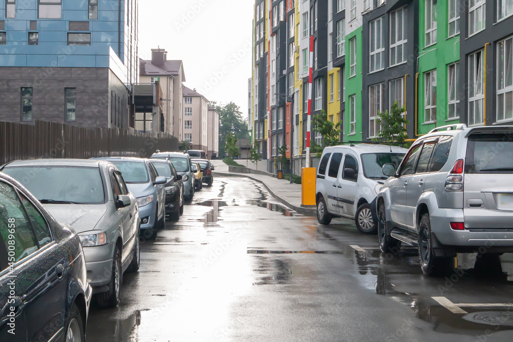 Obraz premium Street in a city without people with parked cars in rainy weather. Rain on the road. Rain and cars. Background of parked cars on a rainy city street. Symmetrical parked cars.