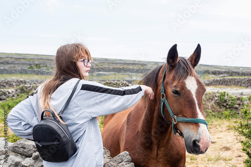 Teenager girl with back pack and brown horse behind dry stone fence. Inishmore, Aran Islands, County Galway, Ireland.