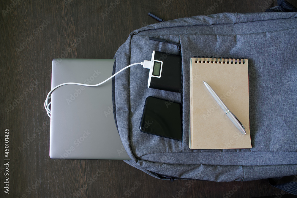 Modern laptop backpack lies with an external battery, smartphone, notepad, pen and silver laptop on a wooden table. The concept of using and recharging gadgets while studying and traveling