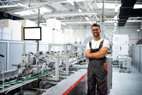 Portrait of an experienced worker in working uniform standing next to modern packaging machine in printing factory. photo