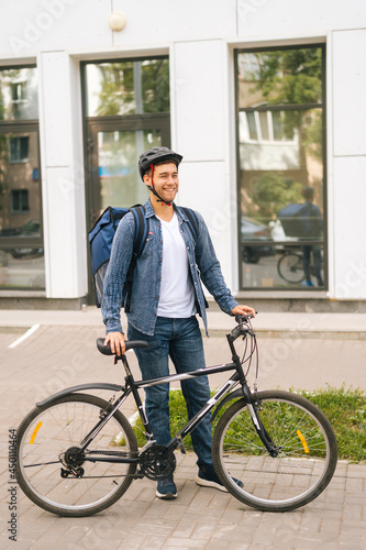 Full length vertical portrait of laughing handsome young delivery man in helmet posing standing near bicycle in city street, looking away. Courier male with thermo backpack delivery food to client
