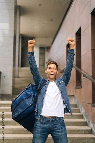 Full length vertical portrait of cheerful handsome young male of food delivery service with large thermal backpack raising hands, celebrating victory standing on stairs, blurred background.