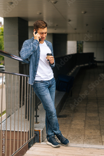 Full length vertical portrait of successful young man talking on mobile phone, standing on balcony terrace with coffee coffee cup leaning on railing of office building, blurred background.