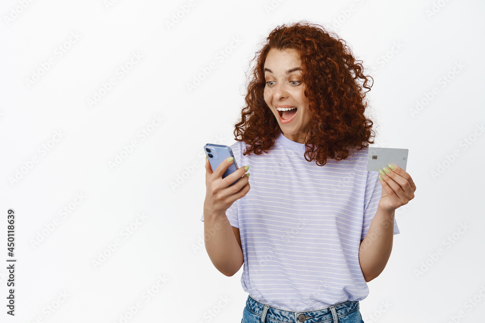 Happy winner looks at mobile phone. Excited redhead girl scream of joy, looking at smartphone screen, holding credit card, shopping online, white background Happy winner looks at mobile phone. Excited redhead girl scream of joy, looking at smartphone screen, holding credit card, shopping online, white background
