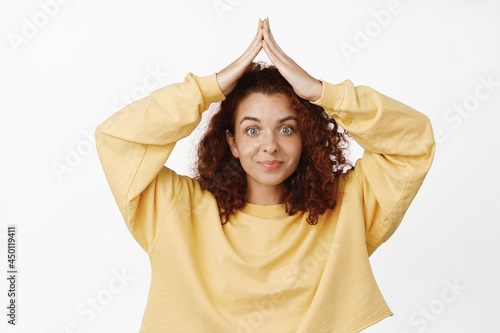 Cute and silly redhead woman making small house rooftop gesture above head, hand roof on top and smiling, standing over white background © Cookie Studio