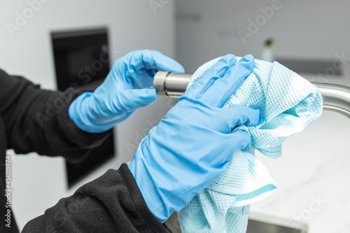 Hands in protective glove cleaning a modern tap with rag of a minimalist domestic kitchen