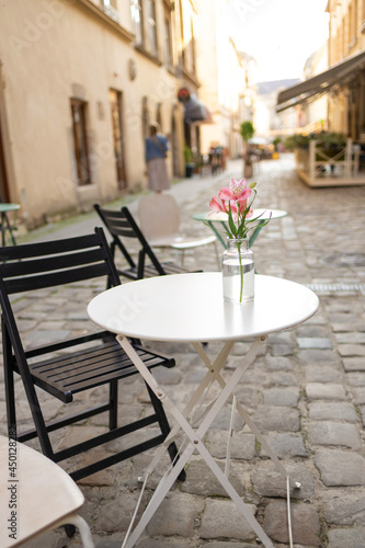 Street view with cafe terrace in the morning in the city. Beautiful delicate pink flower in a transparent glass vase on a white street table