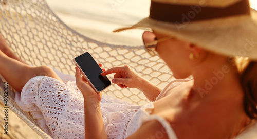 Working at sunset. Young happy female using smartphone while lying in the hammock on the beach