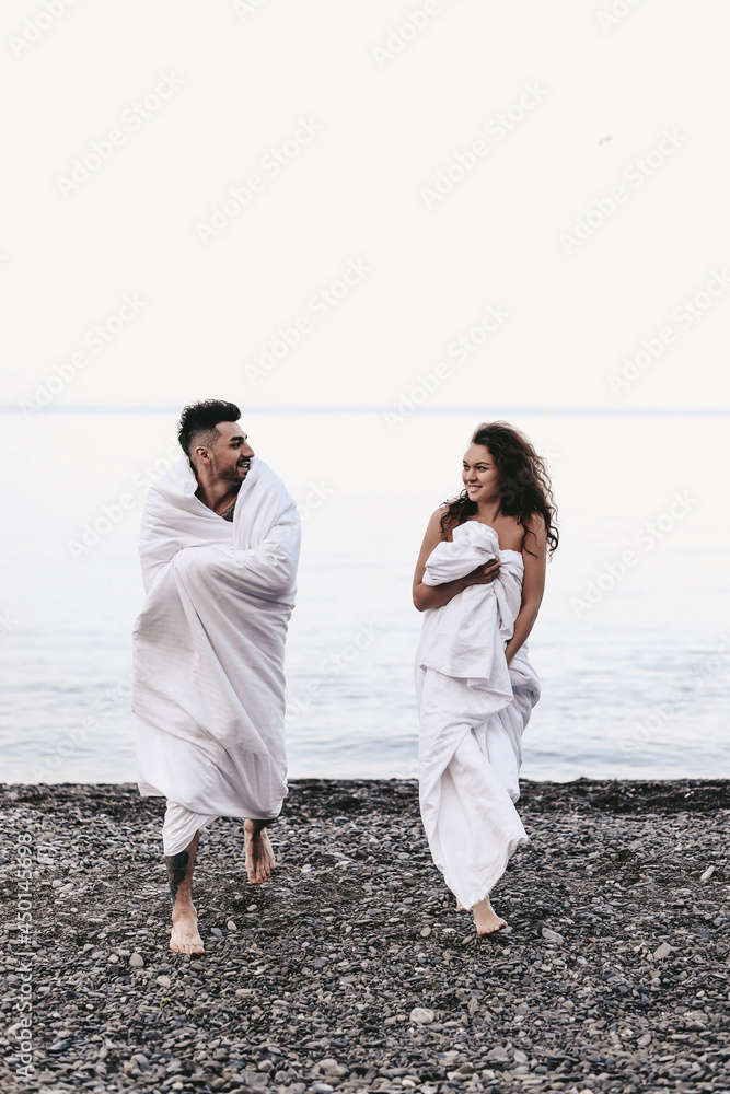 A couple in love are walking together hugging spend the night covered with a white blanket on the ocean beach on a romantic honeymoon in nature. Passionate man and woman of mixed diverse races A couple in love are walking together hugging spend the night covered with a white blanket on the ocean beach on a romantic honeymoon in nature. Passionate man and woman of mixed diverse races