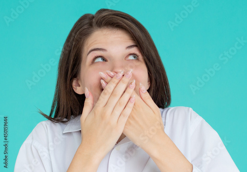 Shocked amazed laughing young brunette woman wearing casual basic white shirt standing covering mouth with hands isolated on pastel blue colour background studio portrait