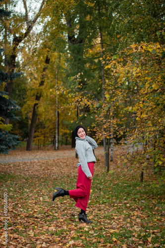 A fashionable Muslim woman stands in the park in a gray sweater and a red skirt. Autumn park  and beautiful yellow leaves.