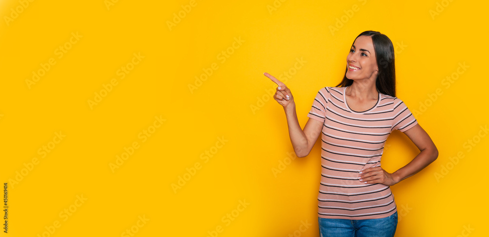 Cute excited smiling young brunette girl is pointing away for some advertisement while stands on yellow wall background Cute excited smiling young brunette girl is pointing away for some advertisement while stands on yellow wall background
