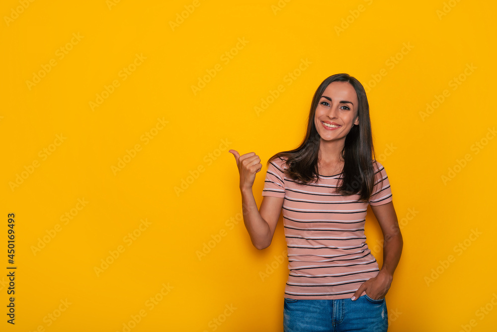 Cute excited smiling young brunette girl is pointing away for some advertisement while stands on yellow wall background Cute excited smiling young brunette girl is pointing away for some advertisement while stands on yellow wall background