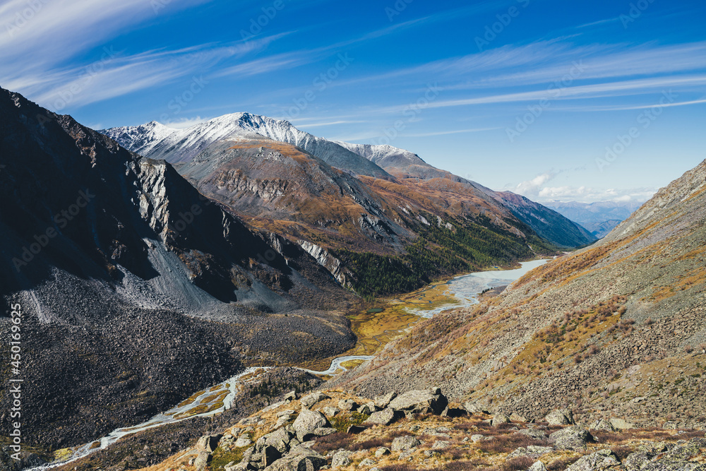 Fototapeta premium Picturesque highland landscape with mountain river and lake in valley among high mountains with snow in autumn colors in sunshine. Awesome alpine view to motley mountain valley in sunlight in fall.