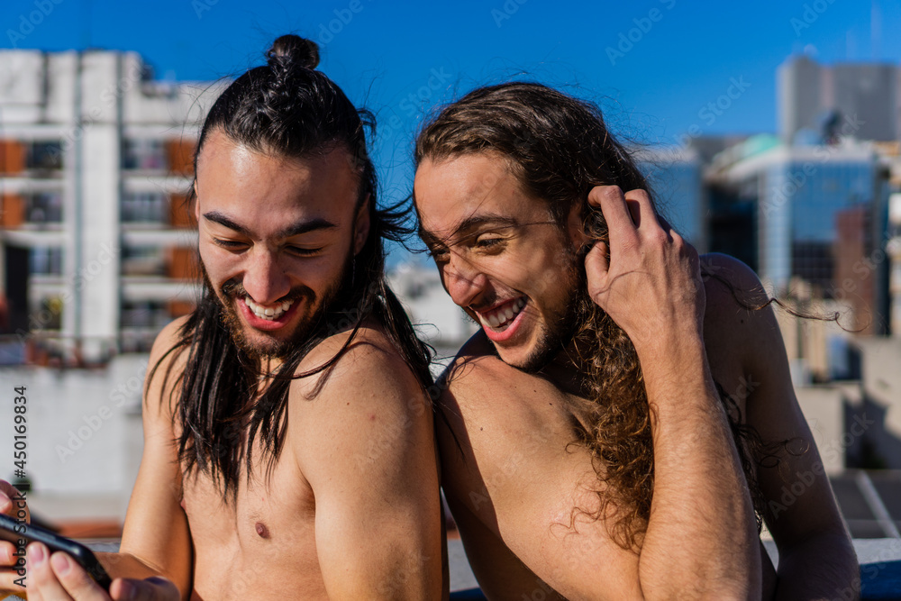 gay homosexual couple of a latin hispanic man and a brazilian man. long haired man laughing sunbathing on the terrace overlooking the city, shirtless looking at his mobile phone.