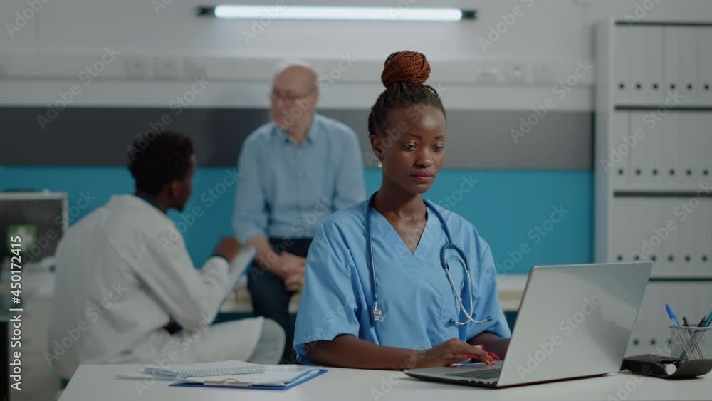 Portrait of nurse typing on laptop keyboard in medical office at facility. Assistant woman with uniform sitting at desk using gadget with technology. Doctor and old patient in background Portrait of nurse typing on laptop keyboard in medical office at facility. Assistant woman with uniform sitting at desk using gadget with technology. Doctor and old patient in background