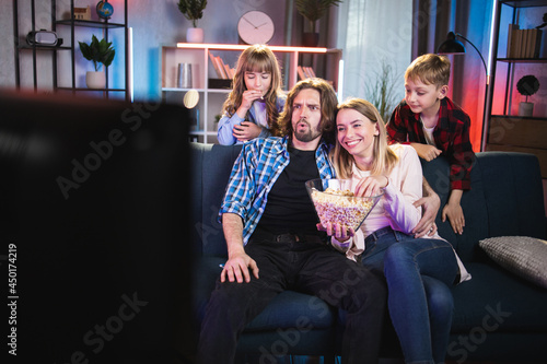 Happy caucasian family of four eating popcorn and watching television while resting together on couch during evening time. Concept of people  lifestyles and leisure.