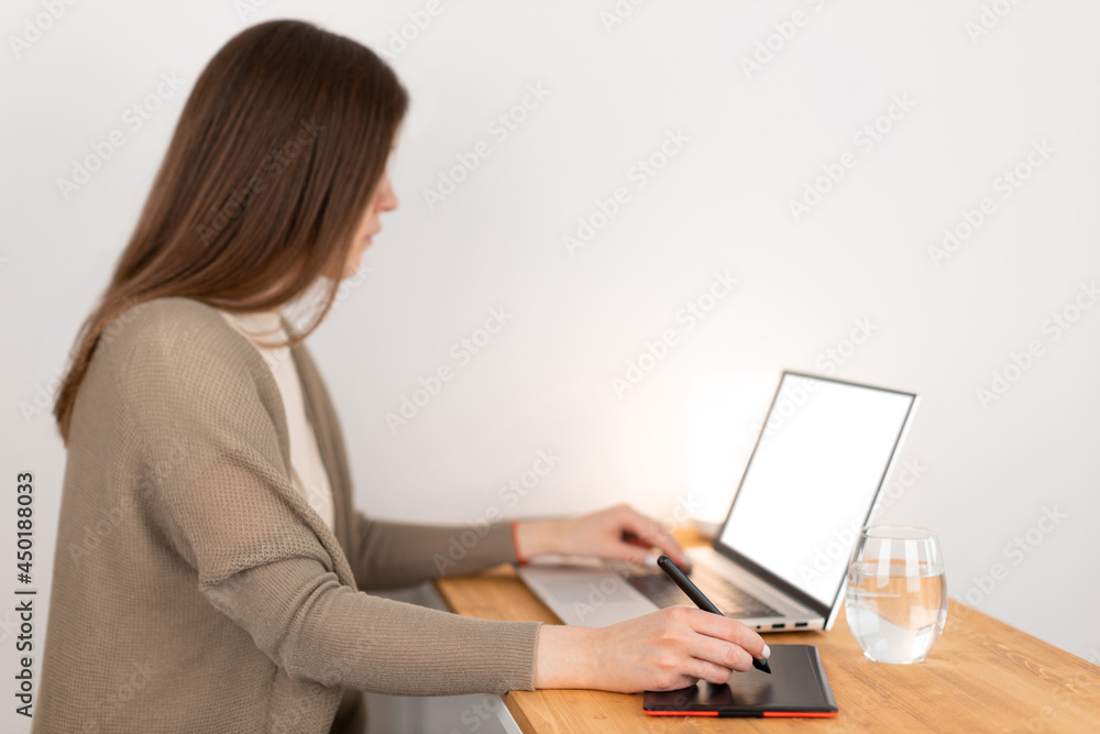 Beauty caucasian girl in beige t-shirt and grey jacket sitting at wooden table opposite laptop and drawing picture on graphic tablet. Designer remotely work from home office. Selective focus on hand. Beauty caucasian girl in beige t-shirt and grey jacket sitting at wooden table opposite laptop and drawing picture on graphic tablet. Designer remotely work from home office. Selective focus on hand.