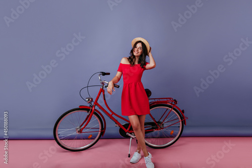 Fototapeta Naklejka Na Ścianę i Meble -  Slim girl in short dress posing with bicycle in studio. Laughing cheerful lady in hat standing beside bike on purple background.