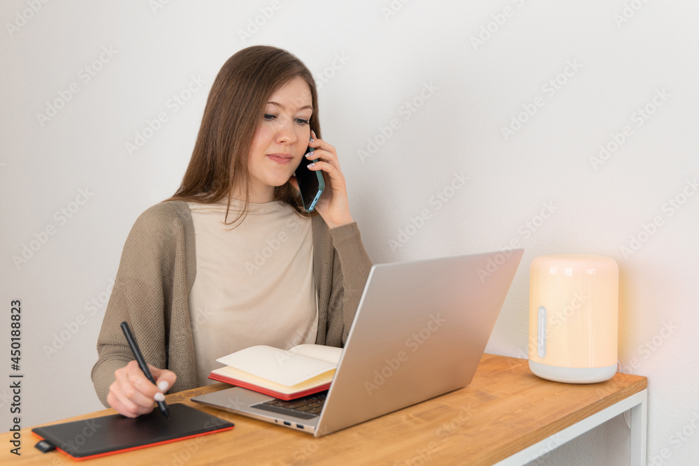 Handsome caucasian woman in beige t-shirt and grey cardigan calling on mobile phone and looking into laptop display. Attractive girl makes handwritten notes on a graphic tablet. Home office concept. Handsome caucasian woman in beige t-shirt and grey cardigan calling on mobile phone and looking into laptop display. Attractive girl makes handwritten notes on a graphic tablet. Home office concept.