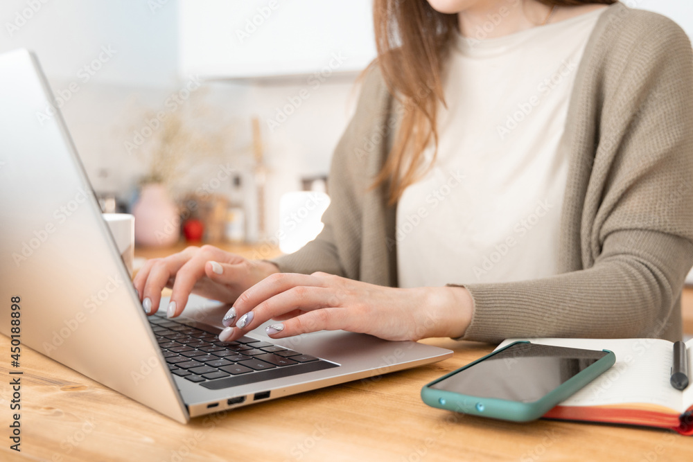 Fototapeta premium Unrecognizable caucasian girl in beige t-shirt and grey jacket typing on computer. On wooden table laptop, smartphone and notepad. Female remotely working from home. Close up, selective focus on hand.