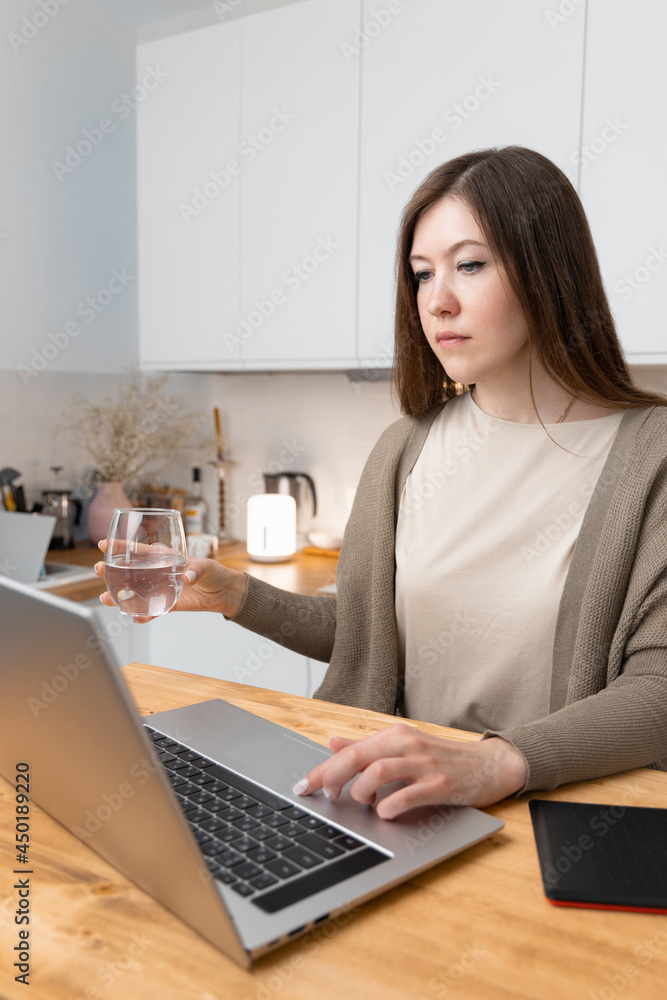 Attractive caucasian female in beige t-shirt and grey jacket took a break from work for a drinking water. At the table laptop and notepad. Home office concept. Woman shopping online or surfing in web. Attractive caucasian female in beige t-shirt and grey jacket took a break from work for a drinking water. At the table laptop and notepad. Home office concept. Woman shopping online or surfing in web.