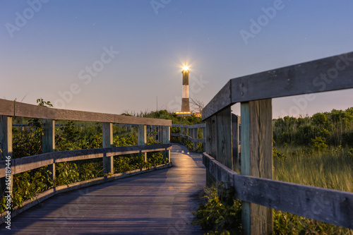 Wooden boardwalk trail illuminated by a tall lighthouse beacon at night. Fire Island, New York