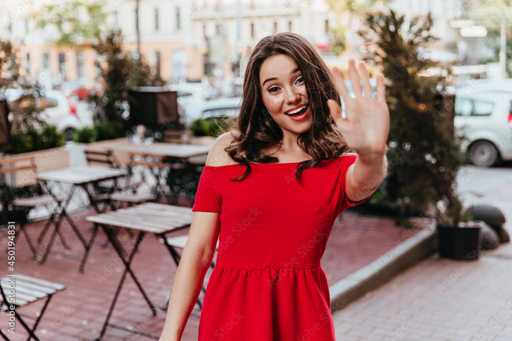 Carefree stylish woman waving hand to camera. Outdoor portrait of cute brunette girl in red dress standing near street cafe.