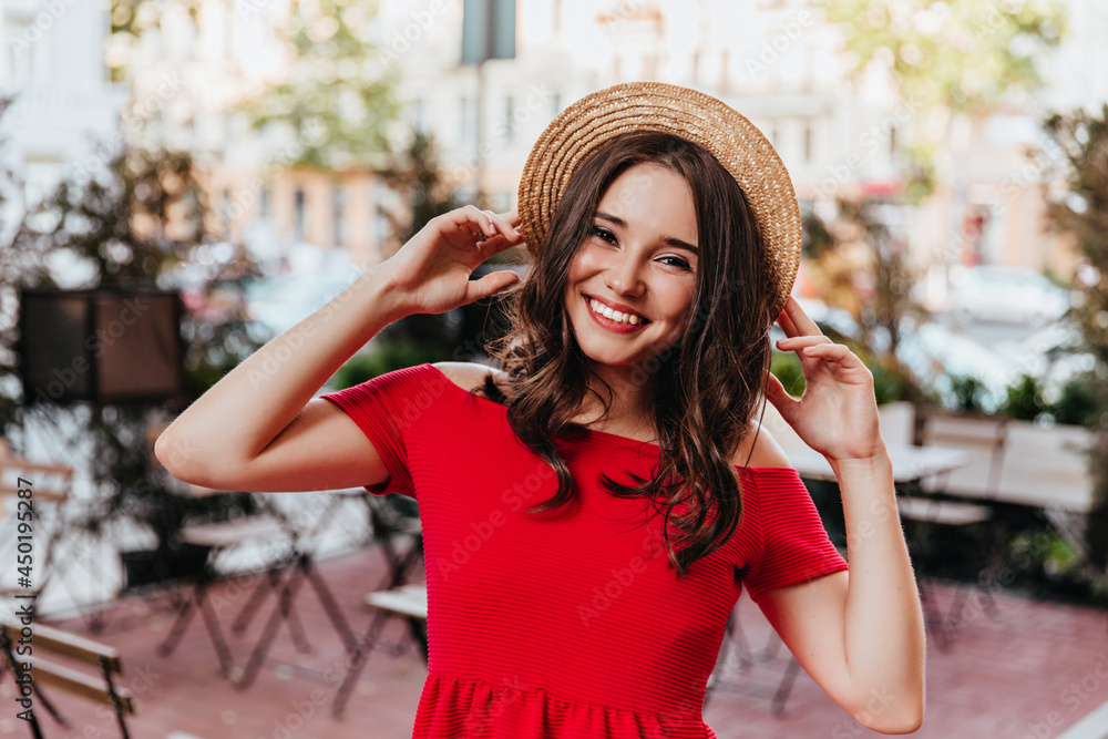 Romantic brown-haired female model posing in straw hat. Outdoor photo of good-humoured girl in red dress standing on city background.