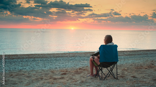 Woman admiring sunset on the sea sitting in folding tourist chair. Relaxing and enjoying nature.