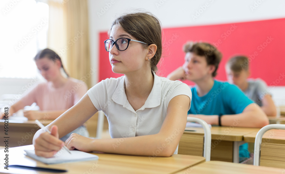 Teen girl listening to lecturer and writing in notebook in classroom