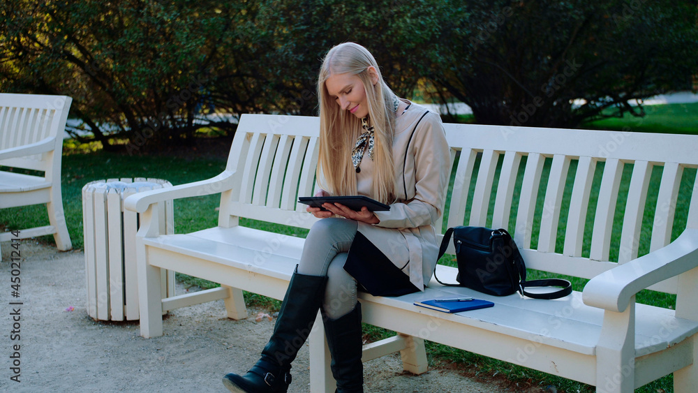 Fototapeta premium Young Beautiful Woman Sits On The Bench In Autumn Park And Works On The Tablet.