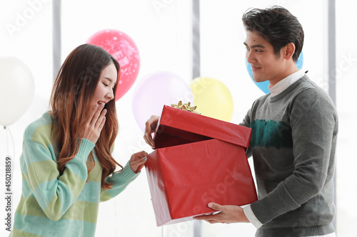 Pretty young Asian woman excited on surprised gift inside red box opening by beloved fiance at romantic indoor party decorated by fancy balloon to celebrate ecstatic love