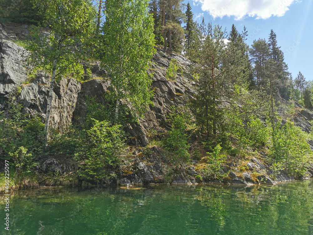 View of the rocky shores and the emerald water of the Marble Canyon in the Ruskeala Mountain Park on a sunny summer day. View of the rocky shores and the emerald water of the Marble Canyon in the Ruskeala Mountain Park on a sunny summer day.