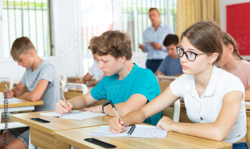 Diligent teenage girl with glasses studying in college with classmates, making notes of teacher lecture
