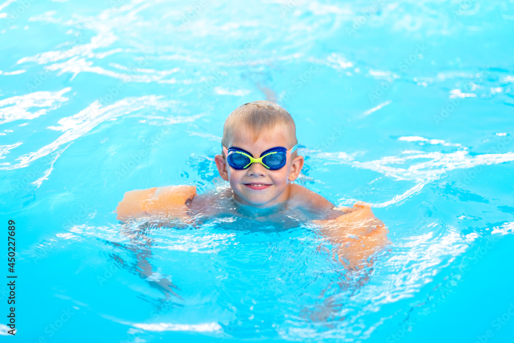 Naklejka premium Portrait smiling boy in swimming pool, child in swimming glasses and inflatable sleeves. Summer travel hotel vacation or classes
