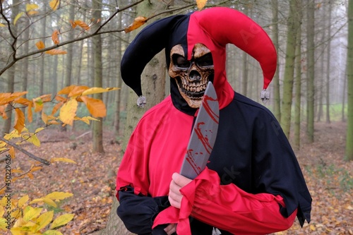  Halloween holiday.scary harlequin. evil jester with a knife in the autumn misty forest.Evil scary jester costume and skull mask.Fear and horror concept. creepy clown in the forest. High quality photo