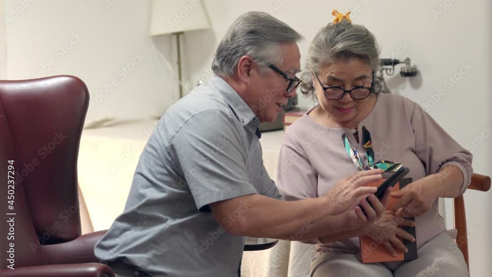 Asian elderly family relaxing together at home. Senior couple using smartphone reading e-book or social media. Retirement family husband and wife enjoy home leisure activity lifestyle with happiness Asian elderly family relaxing together at home. Senior couple using smartphone reading e-book or social media. Retirement family husband and wife enjoy home leisure activity lifestyle with happiness