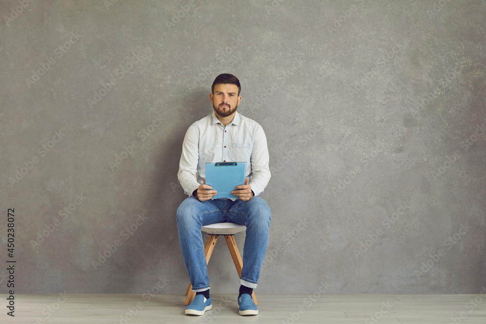 The only job candidate sitting on chair, holding clipboard with resume and waiting for his interview. Unemployed young man in casual shirt and jeans with CV in hands sitting near grey corridor wall