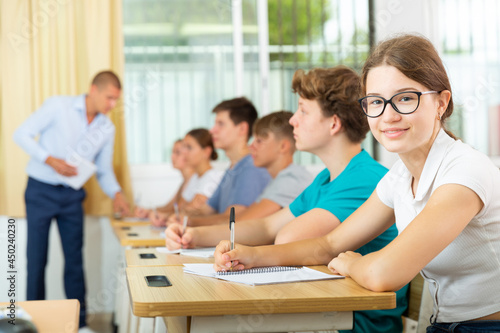 Attractive cheerful teen girl in glasses studying in classroom  listening to lecturer and writing in notebook..