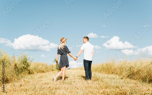 Young couple in love is walking in field holding hands  man and young woman in straw hat and summer dress are looking at each other against blue sky. Romantic walk  trip. Love story concept