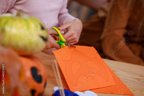 Preparing for Halloween. Close up of a girl's hands cutting out a bat shaped mask on orange paper. Girl at the table with Halloween pumpkins, the foreground is blurred.