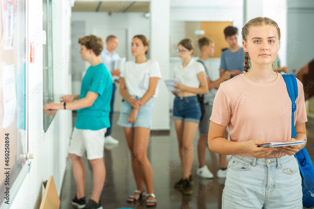 Fototapeta premium Teenage girl standing in school's hallway. Group of young students standing in line in background.