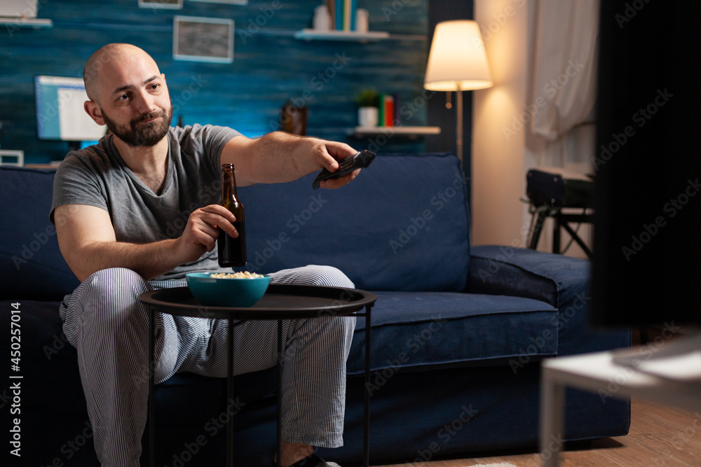 Excited relaxed man spending free time watching TV entertainment series eating popcorn and drinking beer. Funny amused enjoying evening at home sitting on comfortable couch dressed in pajamas Excited relaxed man spending free time watching TV entertainment series eating popcorn and drinking beer. Funny amused enjoying evening at home sitting on comfortable couch dressed in pajamas